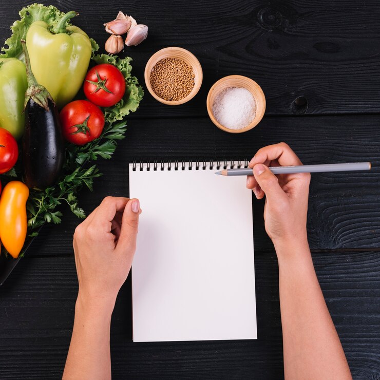 Foto gratuito mano humana con cuaderno y lápiz en espiral cerca de verduras y especias sobre fondo negro de madera