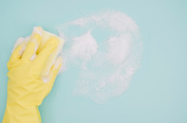 Mano humana con guantes amarillos lavando fondo azul con jabón sud