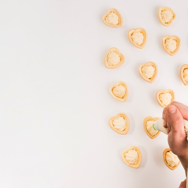 Mano humana poniendo crema fresca en delicioso corazón forma tartaleta