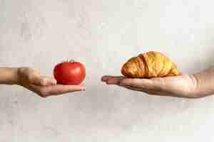 Foto gratuita mano humana que muestra el croissant y el tomate rojo delante de fondo concreto