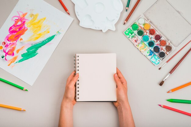 Mano humana que sostiene la libreta espiral con los accesorios de la pintura sobre fondo gris