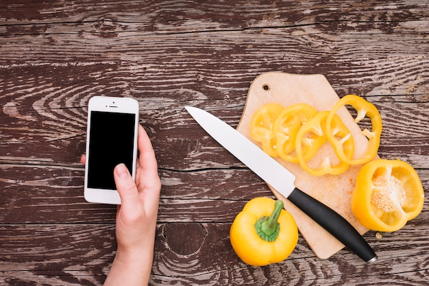 Mano humana sosteniendo teléfono celular con rodajas de pimiento amarillo en tabla de cortar con un cuchillo sobre el escritorio de madera