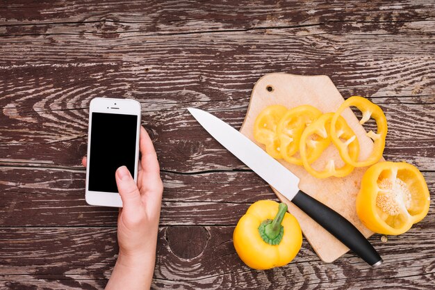 Mano humana sosteniendo teléfono celular con rodajas de pimiento amarillo en tabla de cortar con un cuchillo sobre el escritorio de madera