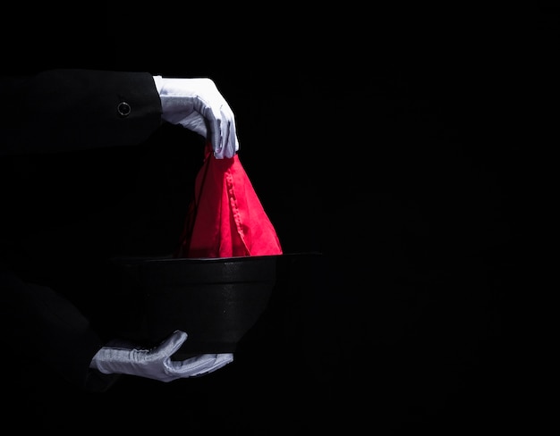 Foto gratuita mano del mago realizando un truco de magia con una servilleta sobre el sombrero negro superior