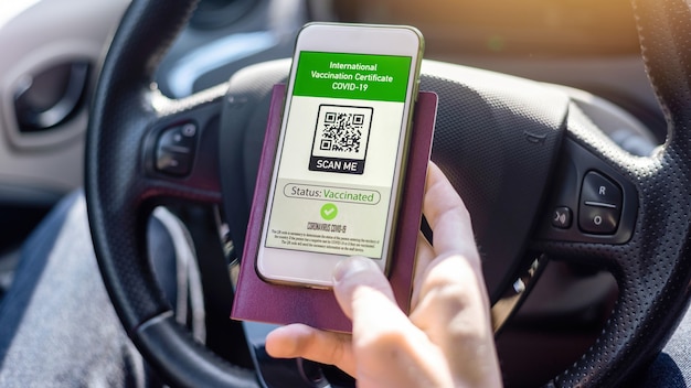 Mano masculina que sostiene el pasaporte y el teléfono inteligente con el código QR del certificado de vacunación internacional COVID-19 en un automóvil