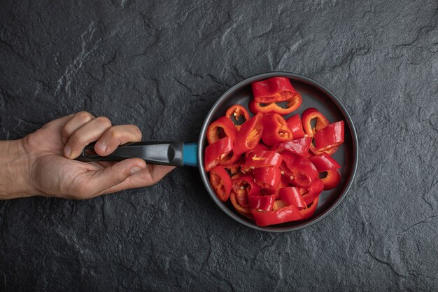 Mano masculina que sostiene la sartén de pimientos rojos en rodajas sobre fondo negro