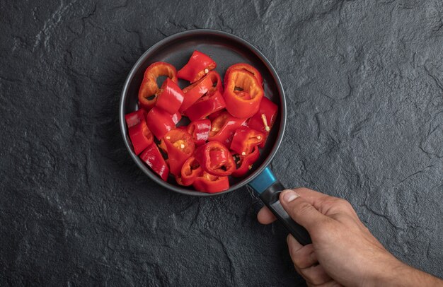 Mano masculina que sostiene la sartén de pimientos rojos en rodajas sobre fondo negro