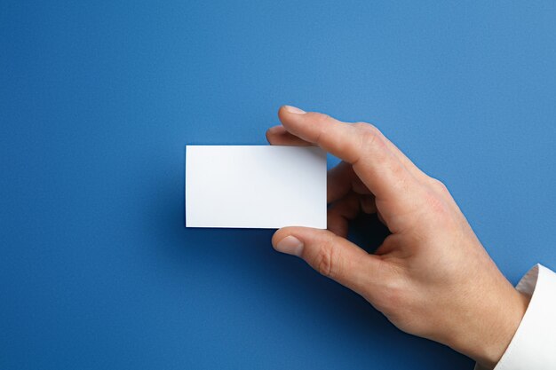 Mano masculina que sostiene una tarjeta de presentación en blanco en la pared azul para texto o diseño. Plantillas de tarjetas de crédito en blanco para contacto o uso en negocios. Oficina de finanzas. Copyspace.