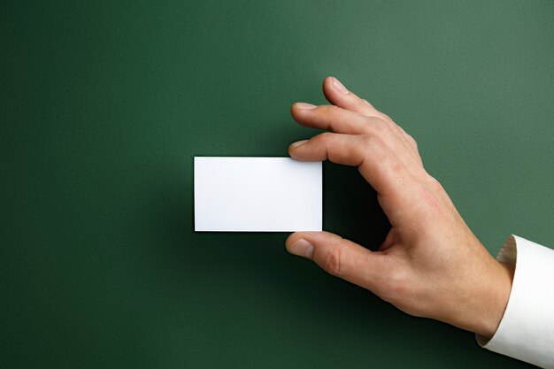 Mano masculina sosteniendo una tarjeta de presentación en blanco en la pared verde para texto o diseño. Plantillas de tarjetas de crédito en blanco para contacto o uso en negocios. Oficina de finanzas. Copyspace.