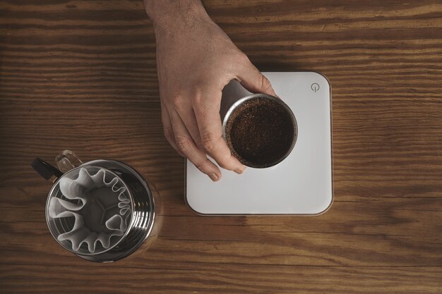 La mano masculina sostiene la taza de plata inoxidable con café molido tostado sobre pesos simples blancos. Cafetera de goteo para café filtrado cerca. Todo sobre la mesa de madera gruesa en la cafetería. Vista superior.