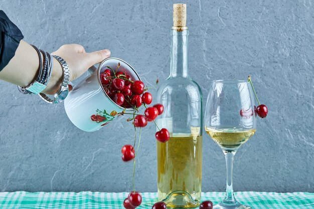 Mano de mujer arrojando una taza de cerezas y una botella de vino blanco con vidrio sobre superficie azul