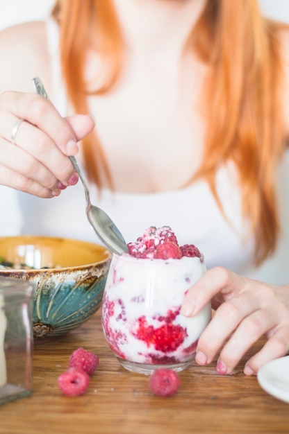 Mano de mujer comiendo frambuesas con yogur en mesa de madera