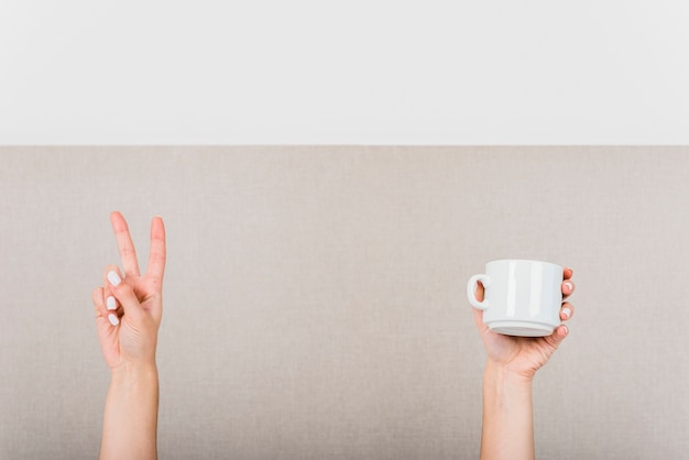 Foto gratuita mano de mujer haciendo señal de victoria y taza blanca contra pared