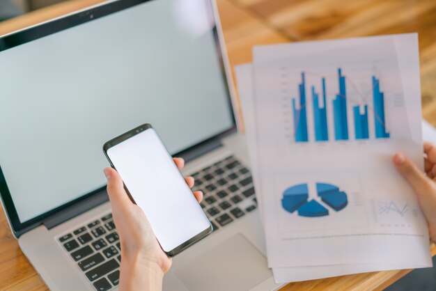 Mano de mujer de negocios con gráficos financieros y el teléfono móvil sobre la computadora portátil en la tabla.