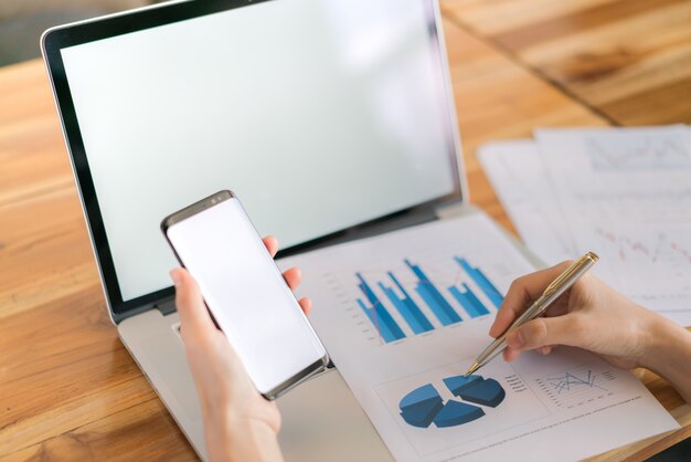 Mano de mujer de negocios con gráficos financieros y el teléfono móvil sobre la computadora portátil en la tabla.