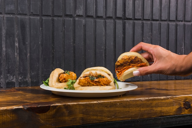 Mano de una mujer con pan de vapor gua bao en plato blanco sobre mesa de madera