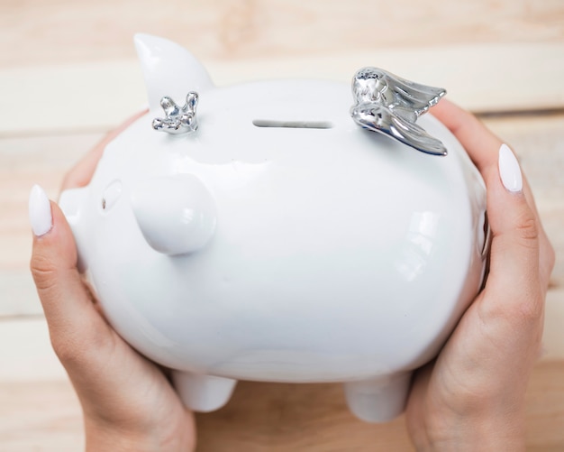 Mano de mujer con piggybank de cerámica blanca con corona de plata y alas