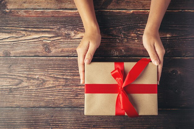 Mano de mujer sosteniendo una caja de regalo con cinta roja sobre un piso de madera