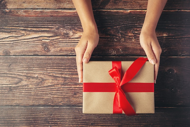 Mano de mujer sosteniendo una caja de regalo con cinta roja sobre un piso de madera