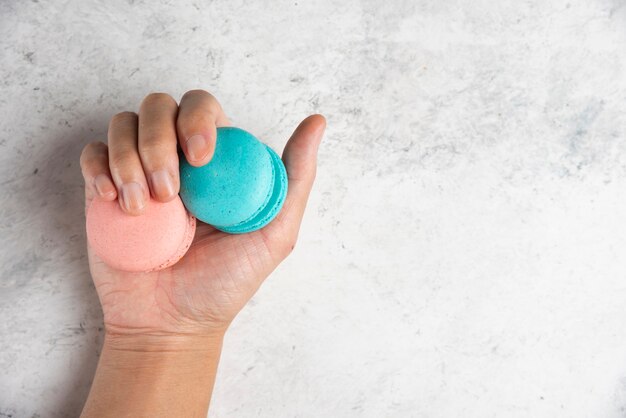 Mano de mujer sosteniendo dos sabrosos macarons sobre fondo de mármol.