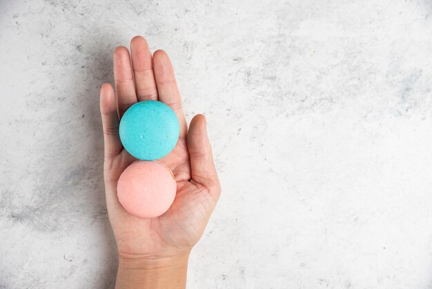 Mano de mujer sosteniendo dos sabrosos macarons sobre fondo de mármol.