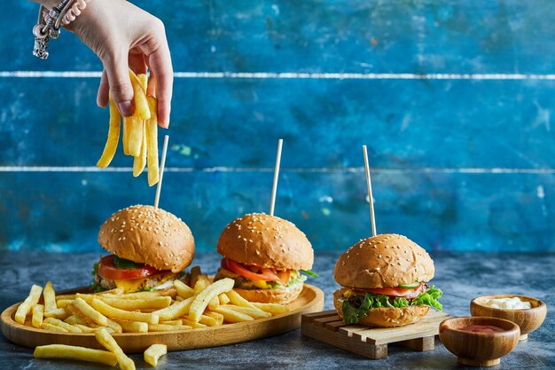 Una mano de mujer sosteniendo patatas fritas con tres hamburguesas con queso, salsa de tomate, mayonesa en placa de madera