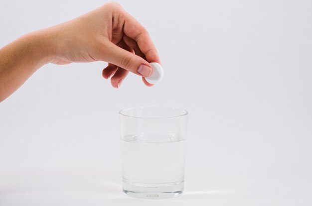 Mano de la mujer sosteniendo la píldora blanca sobre el vaso de agua contra el fondo gris