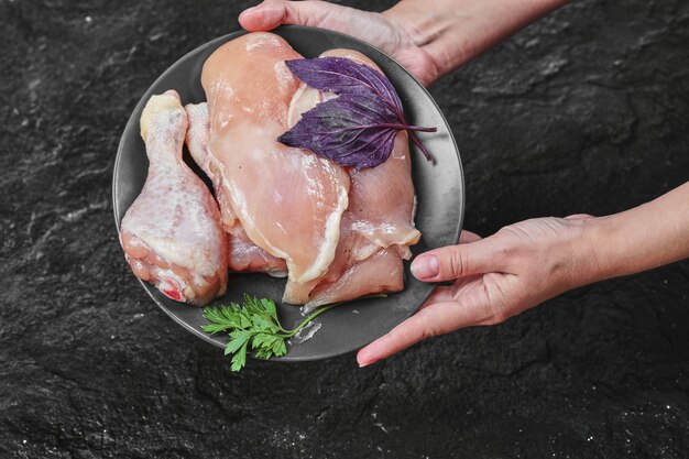 Mano de mujer sosteniendo un plato de partes de pollo crudo con albahaca sobre una superficie oscura