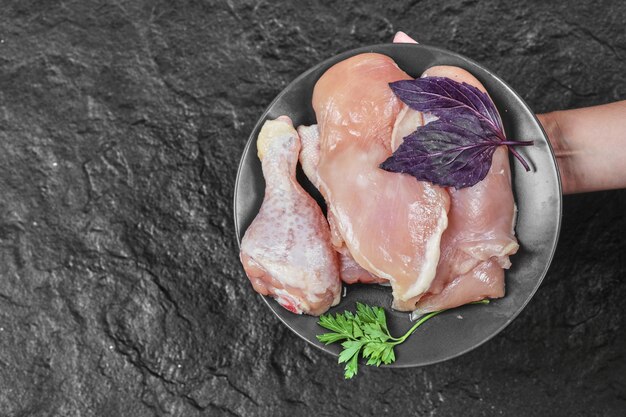 Mano de mujer sosteniendo un plato de partes de pollo crudo con albahaca sobre una superficie oscura
