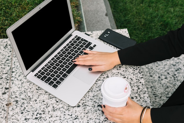 Mano de mujer sosteniendo la taza de café desechable escribiendo en la computadora portátil