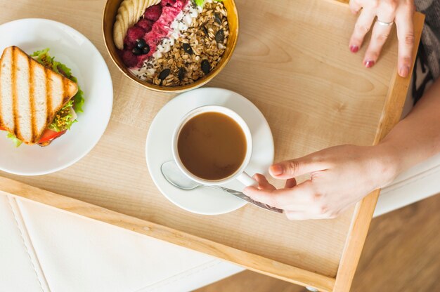 Mano de mujer sosteniendo una taza de té con desayuno saludable en bandeja de madera
