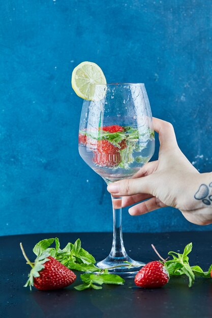 Mano de mujer sosteniendo un vaso de jugo con frutas enteras y menta dentro