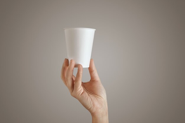 La mano de la mujer sostiene el vaso de papel en blanco vacío para llevar desde la parte inferior, presentación en el centro, vista lateral, aislado, irreconocible