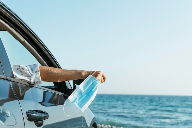 Mano de mujer de tiro medio fuera de la ventana del coche y sosteniendo la mascarilla cerca del mar