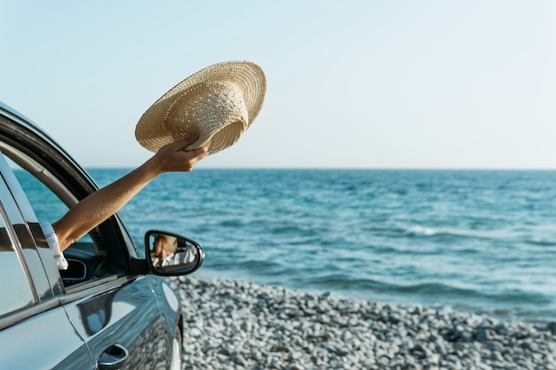 Mano de mujer de tiro medio fuera de la ventana del coche y sosteniendo el sombrero cerca del mar