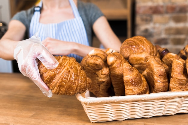 Mano de panadero femenino usando guantes de plástico que toman croissant horneado de la canasta