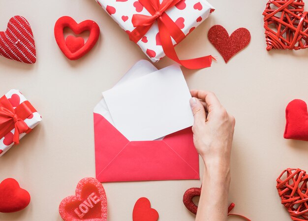 Mano con papel cerca de sobre, regalos y adornos de corazones.