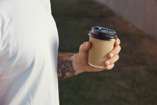 Mano y pecho de un hombre tatuado blanco vestido con camiseta blanca sin etiqueta sosteniendo una taza de café de papel marrón claro
