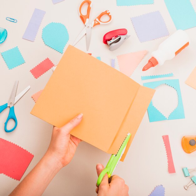 Mano de la persona cortando papel de colores sobre útiles escolares en mesa