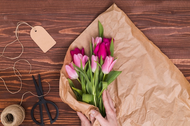 Mano de la persona envolviendo flores de tulipán en papel marrón con una cuerda; cortar con tijeras; etiqueta de precio en el escritorio de madera