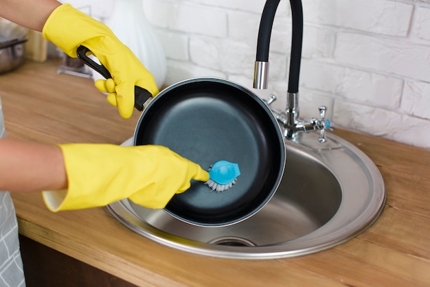 Una mano de persona con guante amarillo lavando sartén con cepillo en la cocina