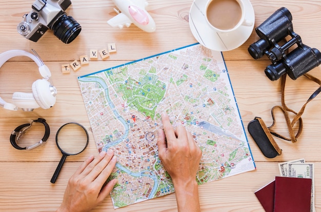 Mano de la persona que apunta a la ubicación en el mapa con una taza de té y accesorios de viajero en superficie de madera