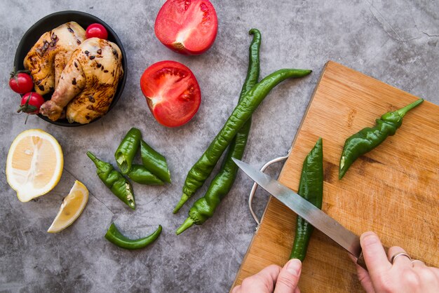 La mano de la persona que corta el chile verde en la tajadera con el pollo asado y los ingredientes en fondo concreto