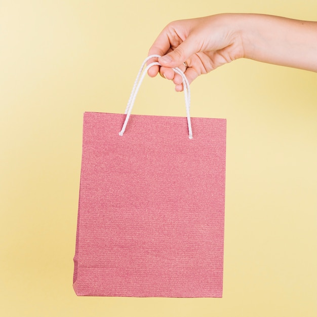 La mano de una persona que sostiene el bolso de compras de papel rosado en fondo amarillo