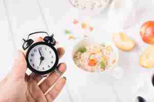 Foto gratuita mano de una persona que sostiene el despertador en mano sobre el desayuno en la mesa