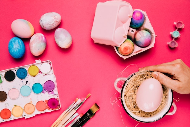 Mano de una persona que sostiene el huevo de Pascua rosado en el pote de cocina con la caja de la acuarela; Pincel y cartón de huevos sobre fondo rosa