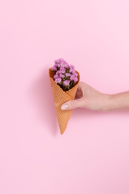 Mano de la persona que sostiene el ramo de flores púrpura en cono de galleta delante de fondo rosa