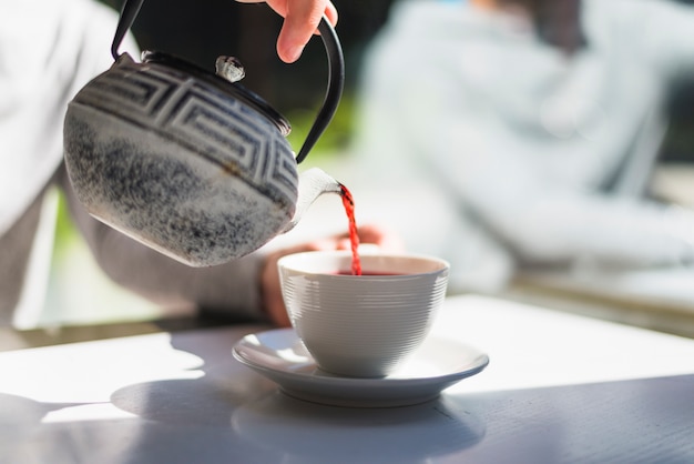 Mano de una persona que vierte té rojo en la taza de cerámica blanca en la mesa blanca en la luz del sol