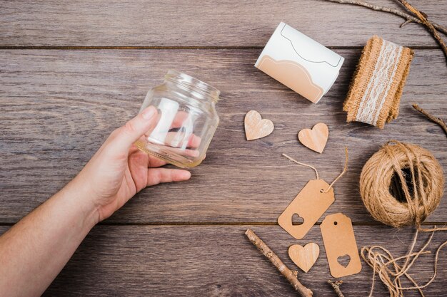 La mano de una persona sosteniendo una botella de vidrio vacía con una cinta de encaje; forma de corazón de madera; Etiquetas e hilo de carrete en la mesa