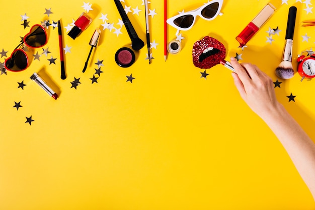 Mano poniendo lápiz labial en broche rojo en forma de labios contra el conjunto de cosméticos.